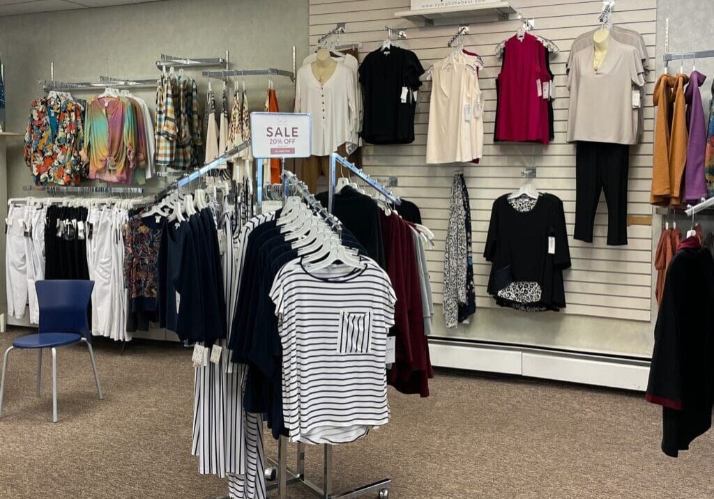 A clothing store display at Bon Jos Boutique features various women's tops and pants on racks and wall hooks. A "SALE 20% OFF" sign sits atop a rack of striped and solid shirts. The store has neutral walls and a blue chair.