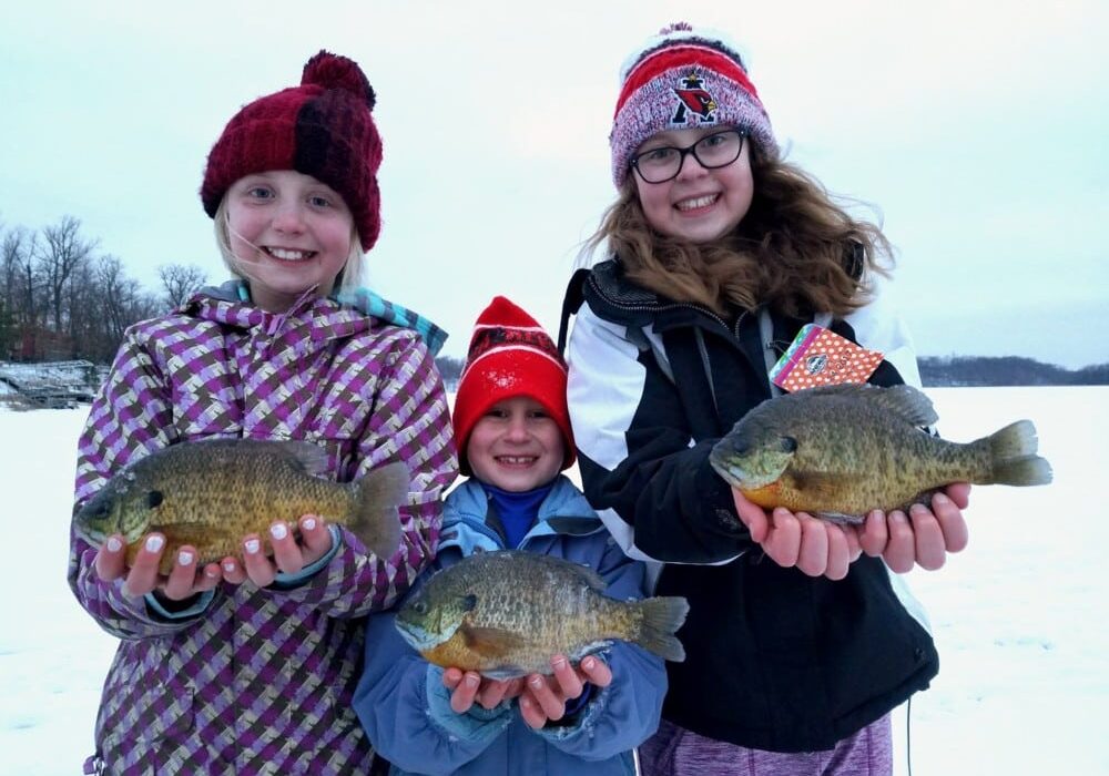 Three smiling children dressed in winter clothing stand on snow holding large fish they caught, with trees and a frozen lake featuring fishable ice in the background.