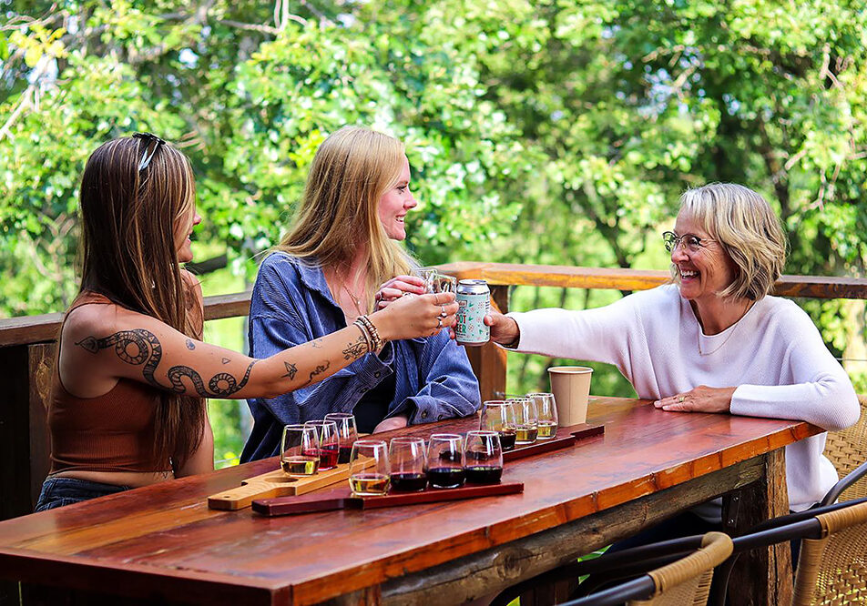 Three women sit at an outdoor wooden table at Burr Vineyards, smiling and toasting drinks together, with flight boards of beverages in front of them and lush green trees in the background.