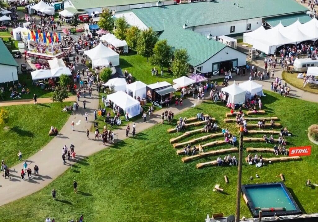Aerial view of the Grape Stomp Festival at Carlos Creek Winery, featuring tents, vendor booths, walking paths, a green-roofed building, and a STIHL-branded woodcutting demonstration area with logs and a small pool on the grass.