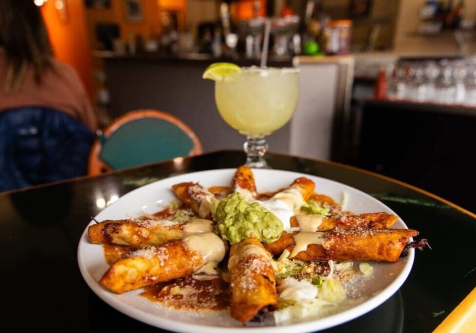 A plate of crispy taquitos topped with sauce, cheese, and a scoop of guacamole sits on a table at Casa Jalisco, with a margarita garnished with a lime wedge in the background at this lively restaurant.