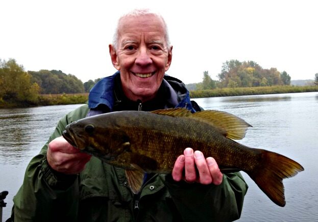 An older man in a green jacket smiles while holding a large fish in front of a river, showcasing his success with fall smallmouth bass fishing tips. Trees and cloudy skies set the scenic background.