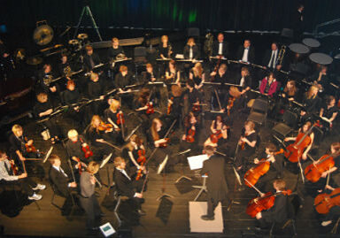 A large orchestra, like the Central Lakes Symphony Orchestra, sits on stage, viewed from above, with musicians playing various instruments. A conductor stands on a podium facing the ensemble—capturing the essence of live music in Alexandria MN.