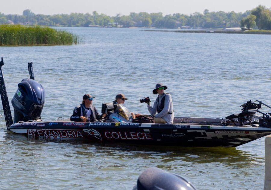 Three fisherman sit in a black Alexandria College fishing boat on a lake, surrounded by water and green shoreline, with fishing equipment visible onboard—ready for their next Champions Tour adventure.