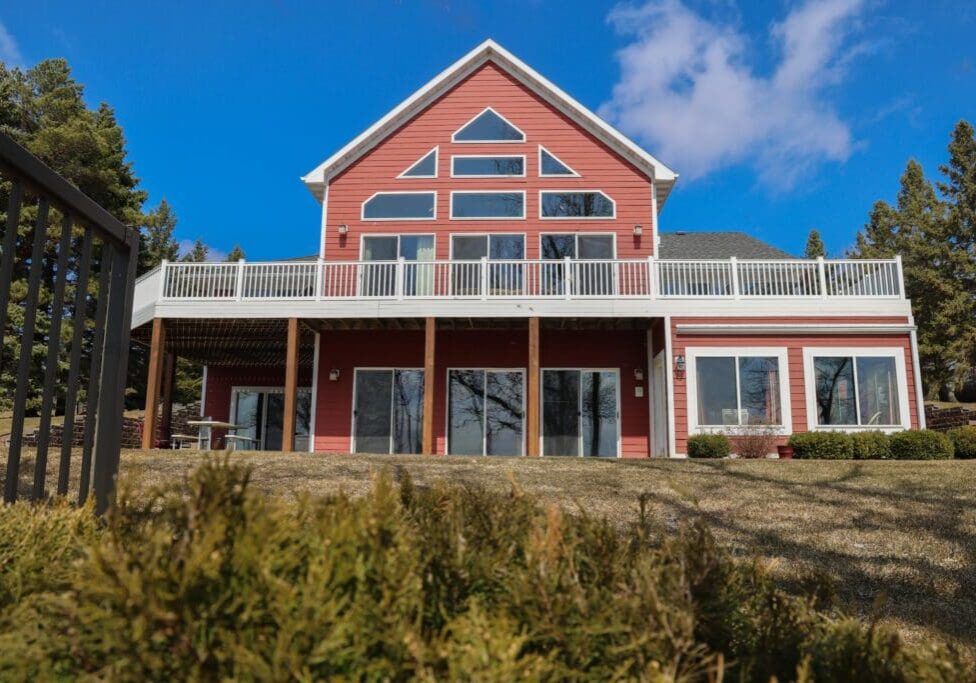 A two-story red chateau with white trim and large windows, featuring a spacious upper deck and a grassy lawn, surrounded by trees under a bright blue sky with scattered clouds.