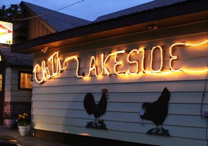 A building at dusk with "CHAIN LAKESIDE INN" spelled in glowing lights on the wall, two chicken silhouettes painted below, and a lit motel sign in the background evoke the charm of Chet's Lakeside Inn.
