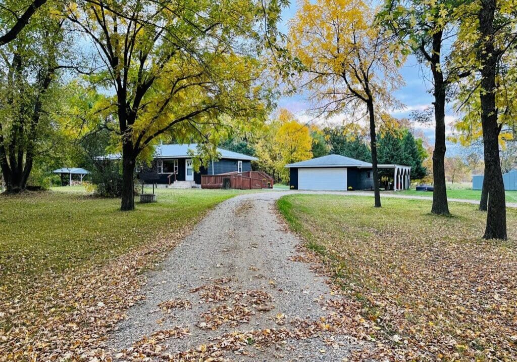 A gravel driveway leads to a one-story Countryside Oasis with a porch and detached garage, surrounded by trees in autumn foliage and a lawn covered in fallen leaves.