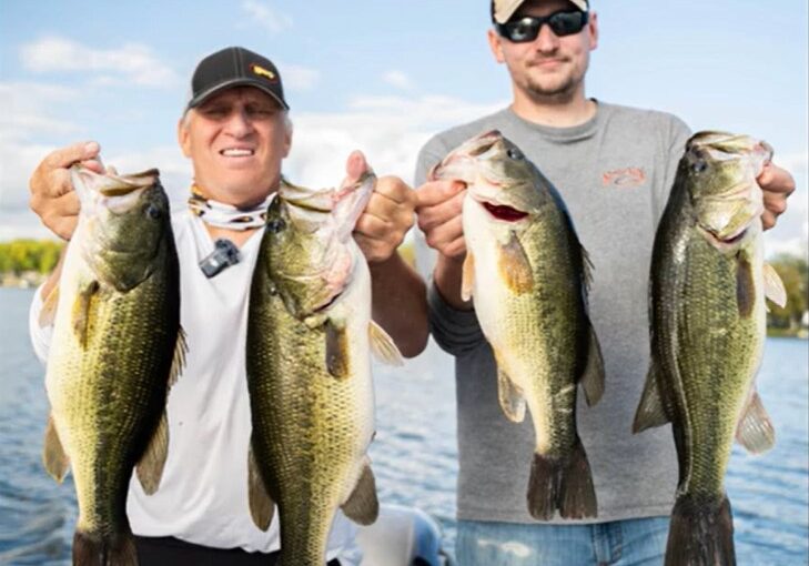 Two men standing outdoors on a boat, each holding up two large bass caught with crankbaits, smiling at the camera, with a lake and blue sky in the background.