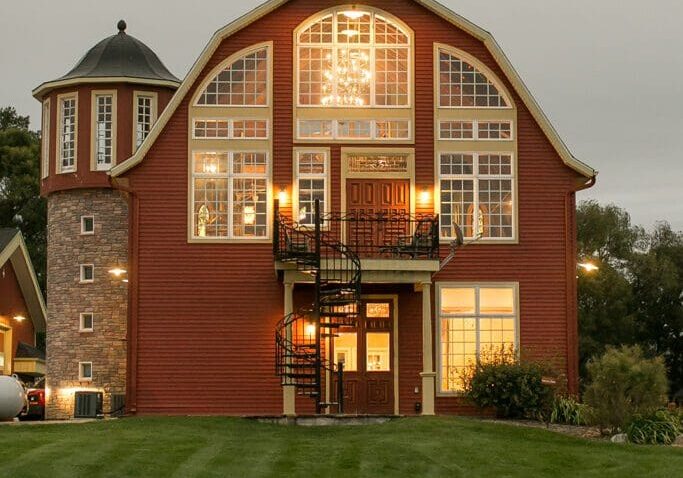 A large red barn-style house, known as Crooked Willow, features numerous large windows warmly lit from inside. It has a round turret on the left and a black spiral staircase at the front, all set on a manicured green lawn under a cloudy sky.