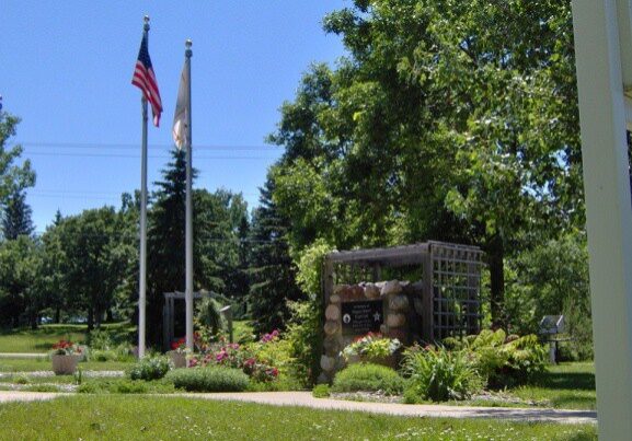 Two flagpoles with American and Illinois state flags stand in Curtis A Felt Memorial Park, a sunny green space with blooming flowers, trees, a stone sign surrounded by plants, and a trellis structure behind the sign.