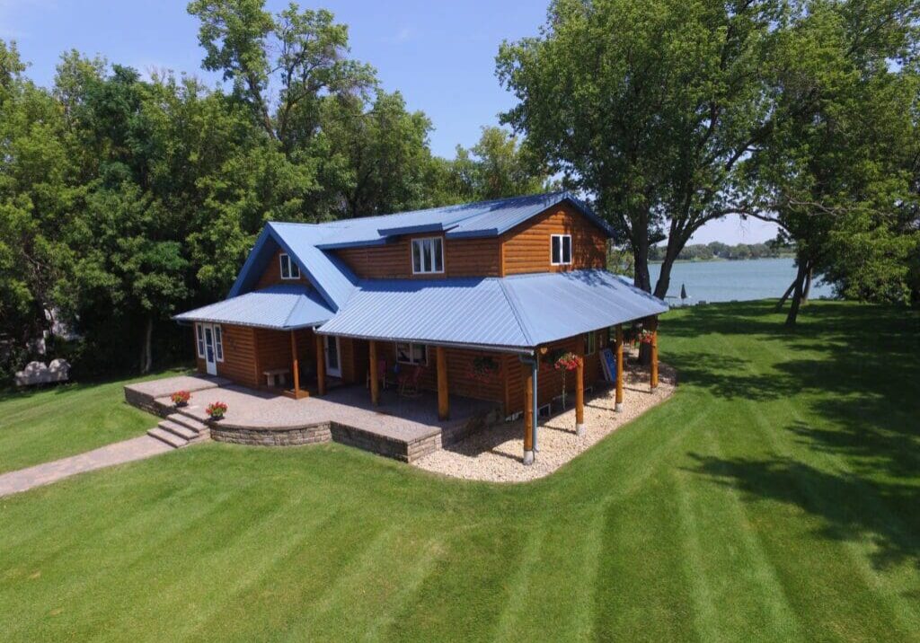 The McCabe Cabin, with its blue metal roof, sits on a well-manicured lawn surrounded by trees. This charming log home features a spacious stone patio and overlooks a serene lake under a clear sky.