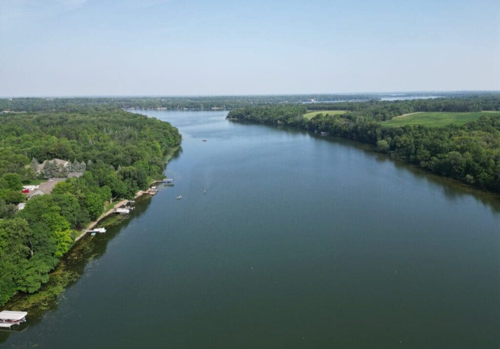 A wide river, reminiscent of Lake Victoria's serene beauty, curves gently through a lush, green landscape with trees lining both banks. Small docks and boats are visible along the left shore under a clear blue sky.