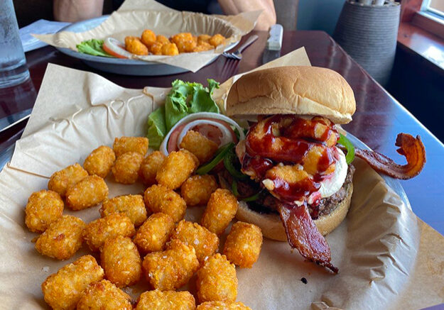 A tray at DJ's Tap House with crispy tater tots, a burger topped with bacon, onion rings, and BBQ sauce, plus lettuce, tomato, and onion on the side. Another similar meal sits in the background on a wooden table.