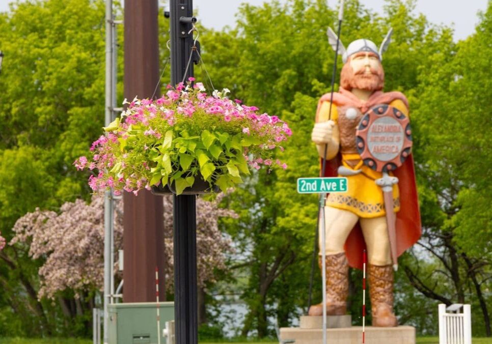 A large Viking statue stands behind a street sign reading "2nd Ave E" and a pole with a hanging basket of pink and green flowers, blooming brightly in May, with lush green trees in the background.