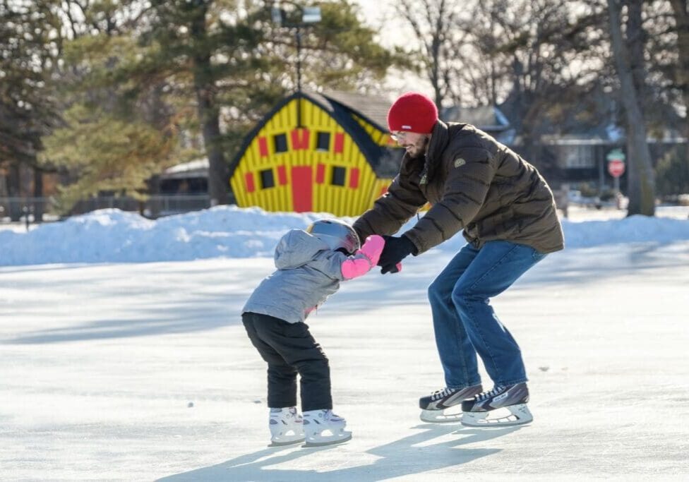 An adult and a child ice skate outdoors at Noonan Park on a sunny winter day. The adult helps the child balance as they glide across the frozen surface, with snow, trees, and a colorful yellow and red shed in the background.