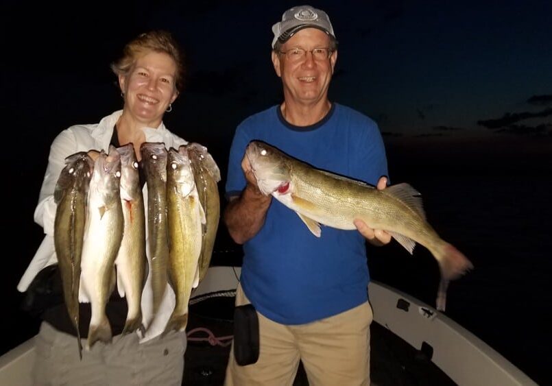 Two smiling people on a boat at night hold up their catch from late August; the person on the left shows off a variety of five fish, while the person on the right displays a single large fish.