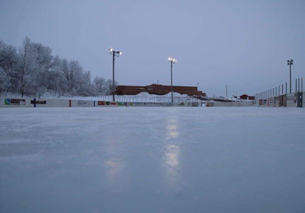 An outdoor ice skating rink at dusk, with snowy trees and Discovery Middle School’s low building in the background. Several tall lights illuminate the rink, reflecting on the smooth ice surface.