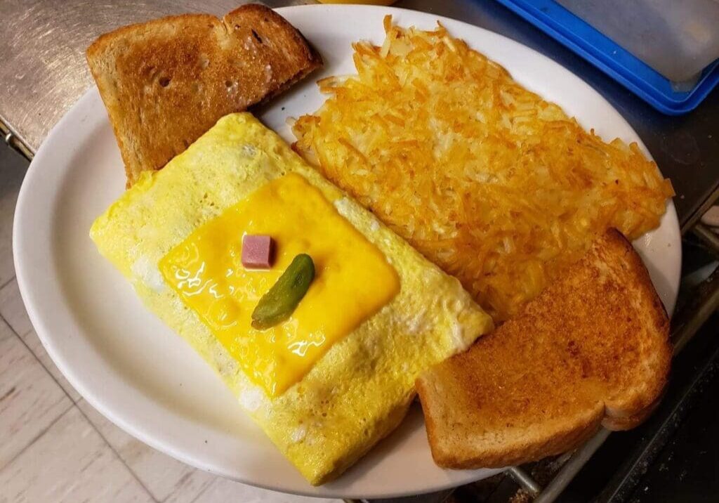 A Downtown Diner breakfast plate featuring two slices of toasted bread, a rectangular omelet topped with melted cheese, diced ham, and a jalapeño slice, served alongside golden hash browns.