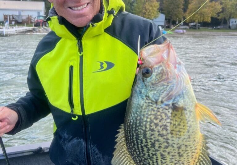 A smiling man in a yellow and black jacket holds up a large crappie fish on a fishing line while fishing the Midwest from his boat on a lake, with trees and houses visible in the background.