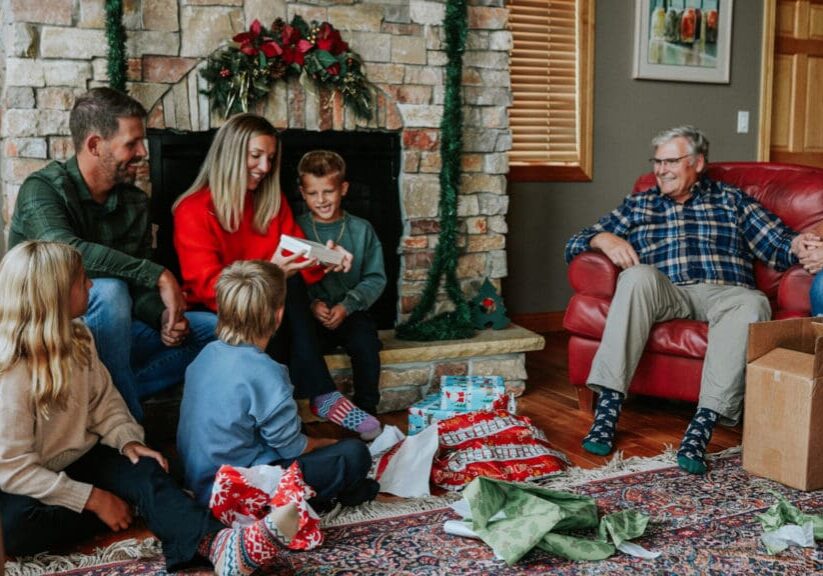 A family sits together in a cozy living room, exchanging and opening gifts in front of a decorated fireplace. As they enjoy the moment, their laughter suggests excitement to explore Alexandria MN after the holiday celebrations.