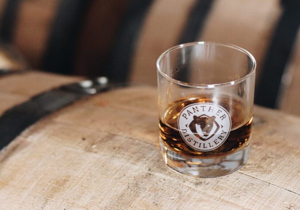 A glass with the Panther Distillery logo, filled with dark liquor, sits on top of a wooden barrel adorned with black metal bands in the background, capturing the authentic spirit of Panther Distillery.