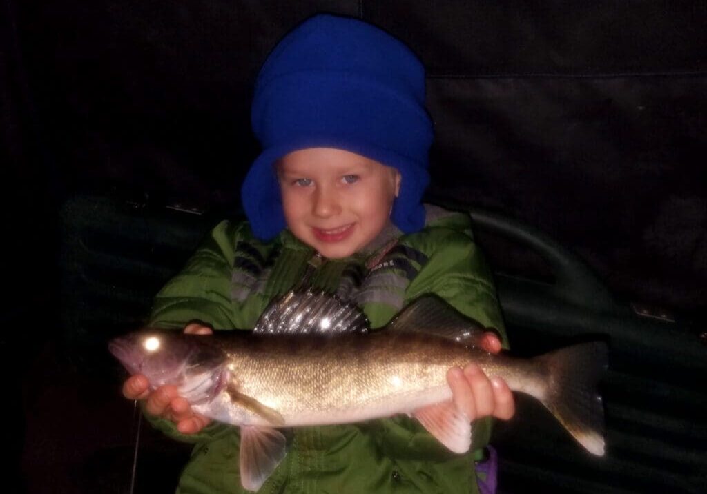 A young child in a blue hat and green jacket smiles while holding a freshly caught fish with both hands. The background is dark, suggesting an outdoor setting—perhaps after an exciting day of ice fishing.