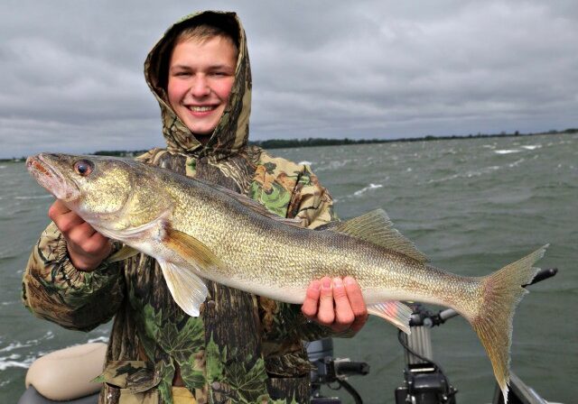 A smiling person in a camouflage jacket holds a large fish on a boat, with choppy water and a cloudy sky in the background—an exciting day fishing for walleye.