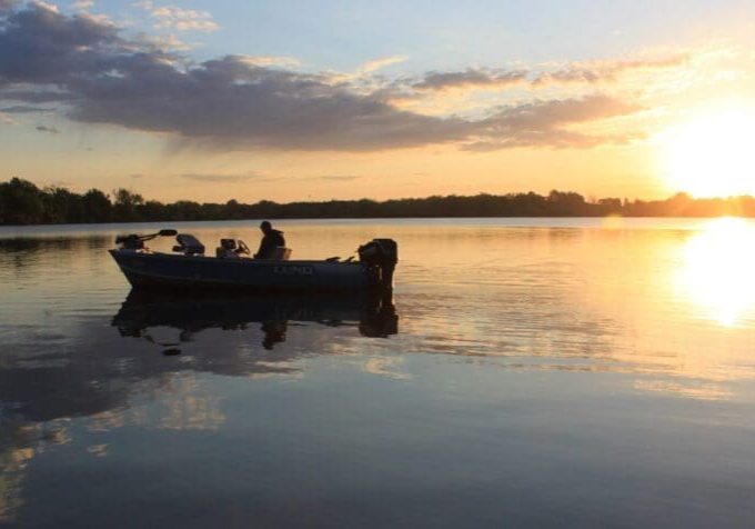 A small boat with two people floats on Lake Agnes at sunset, the sky glowing orange and reflections shimmering on the water. Trees line the horizon in the background.