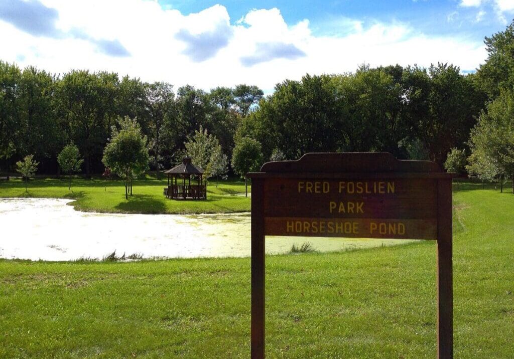A wooden sign reading "Fred Foslien Park, Horseshoe Pond" stands on green grass by a small pond. In the background of Fred Foslien Park, trees and a gazebo are visible under a partly cloudy sky.