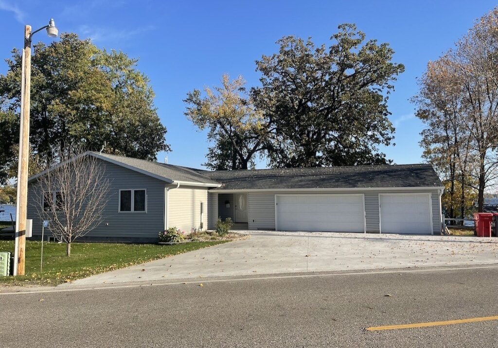 Single-story Private Vacation Home with light-colored siding, a spacious three-car garage, and a wide concrete driveway. The yard features trees, while the house is set against taller trees under a clear blue sky.