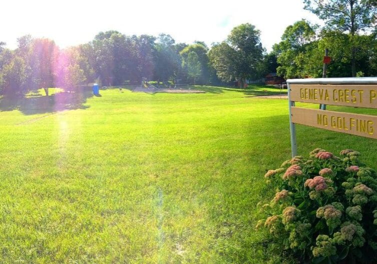 A sunlit park with lush green grass and trees. In the foreground, a wooden sign reads "GENEVA CREST PARK" and "NO GOLFING" beside flowering plants. Geneva Crest Park appears spacious and serene.