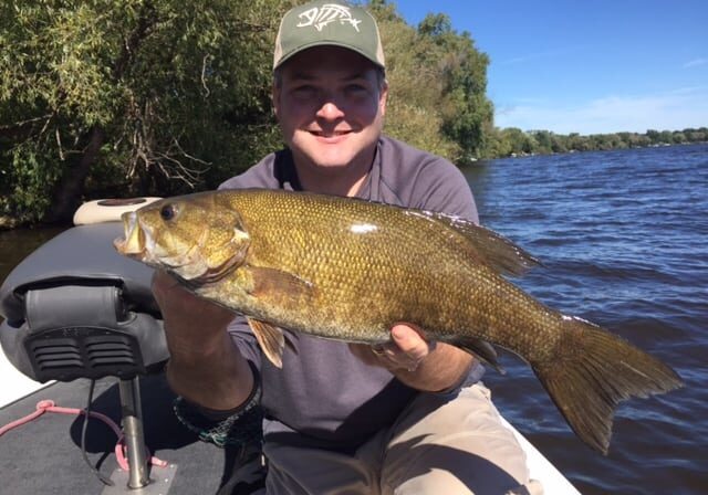 A man in a green hat sits in a boat on a river, smiling as he holds a large bass with both hands—proof of a successful day of bass fishing. Trees and blue sky are visible in the background.