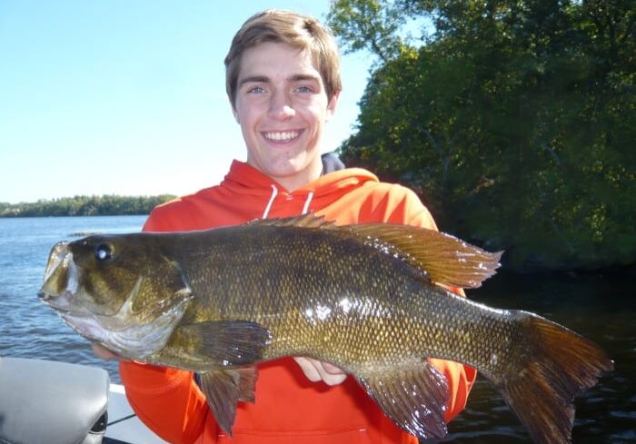 A smiling person in an orange hoodie holds a large fish in front of a river, capturing the excitement of a fall small mouth feeding frenzy, with trees and blue sky in the background.