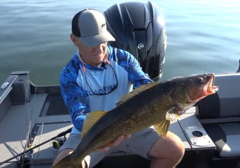 A man in a blue shirt and cap sits in a boat on the water, holding a big walleye with both hands. A fishing rod and boat engine are visible in the background—classic fishing the Midwest style.
