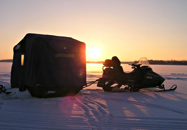 A snowmobile is parked on a snowy landscape at sunset, towing a dark-colored ice fishing shelter—highlighting the excellent ice fishing conditions in Alexandria as long shadows stretch across the gleaming snow.
