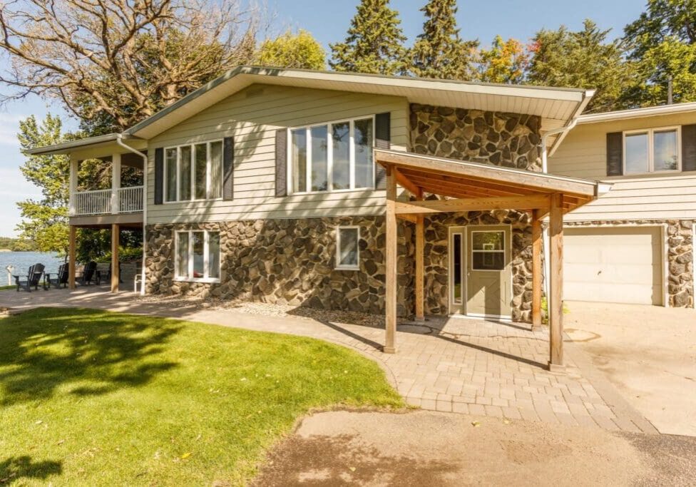 A two-story graystone house with wood siding, large windows, a covered entryway, attached garage, and a patio overlooking a lake, surrounded by trees and green grass under a clear sky.