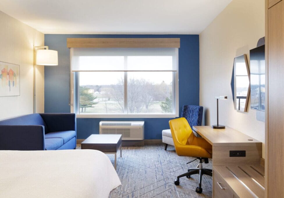 A modern Holiday Inn Express hotel room with a blue accent wall, a sofa, a work desk with a yellow chair, an armchair, and a large window overlooking trees and a grassy area.