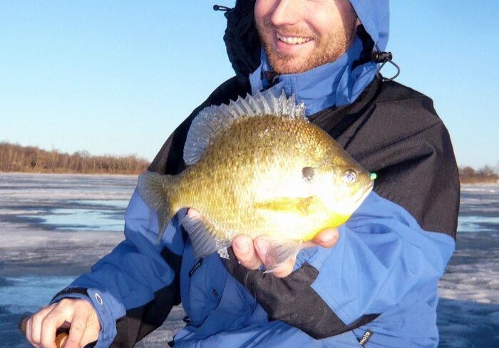 A person in a blue jacket smiles while holding a big panfish in Alexandria, sitting on an icy surface with a fishing rod in hand under a clear blue sky.