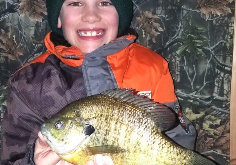 A smiling child in winter clothing and a beanie holds a large sunfish with both hands, proudly posing as if they've just reeled in a walleye. The background features a camouflage-patterned wall.