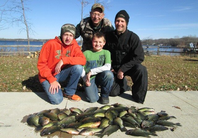Four people kneel and smile behind a large display of caught fish laid on the ground by a lake on one of the season’s First Frozen Lakes, with clear skies, trees, and water in the background.