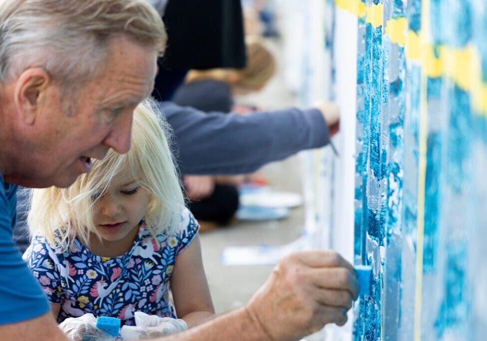 An older man and a young girl with blonde hair paint a mural together outdoors as part of the Alexandria community mural project. Both are focused on applying blue paint to a wall, while others work on the community mural in the background.