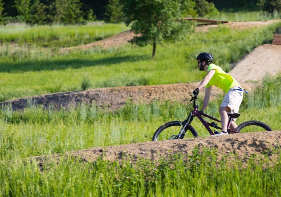 A person wearing a helmet, neon green shirt, and shorts enjoys physical activity as they ride a mountain bike over a dirt trail surrounded by green grass and trees on a sunny day.