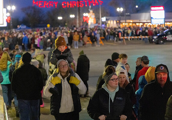 A large crowd gathers outdoors at night under a lit "Merry Christmas" sign during one of the lively Christmas events in Alexandria. In the foreground, a woman carries a child on her shoulders while people in winter clothes enjoy the festive, illuminated scene.