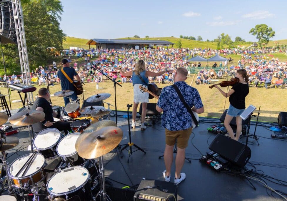 A band performs on an outdoor stage with drums, guitars, and a violin at Summer Sounds by Brophy, facing a large seated crowd on a grassy field under a sunny sky.