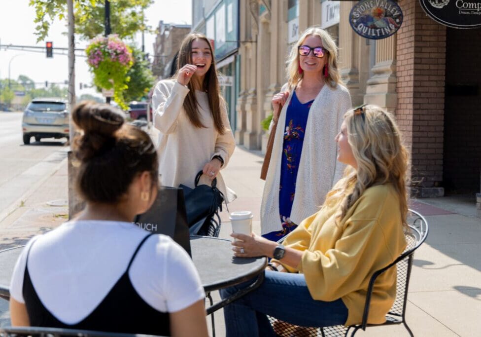 Four women enjoy a sunny day outside at a sidewalk café in downtown Alexandria; two sit at a table with coffee while two stand nearby, smiling and chatting. Cars and shops line the background street.