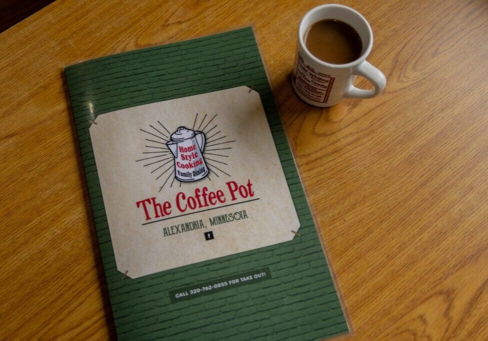 A menu from the Coffee Pot Cafe in Alexandria, Minnesota, lies on a wooden table next to a mug filled with coffee.