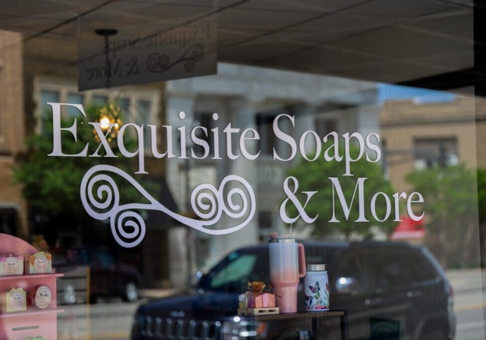 Storefront window with "Exquisite Soaps & More" in white lettering and a decorative swirl design, reflecting the street, nearby buildings, and a parked car—perfectly capturing the charm of Exquisite Soaps.