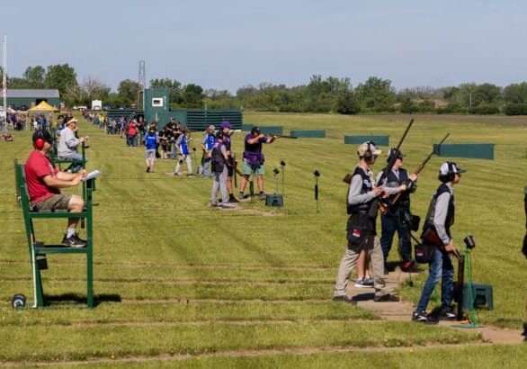 Student athletes participate in the Alexandria Trap Shooting Championship at an outdoor range. Shooters, judges, and spectators line several lanes on a grassy field under sunny skies with trees in the background.