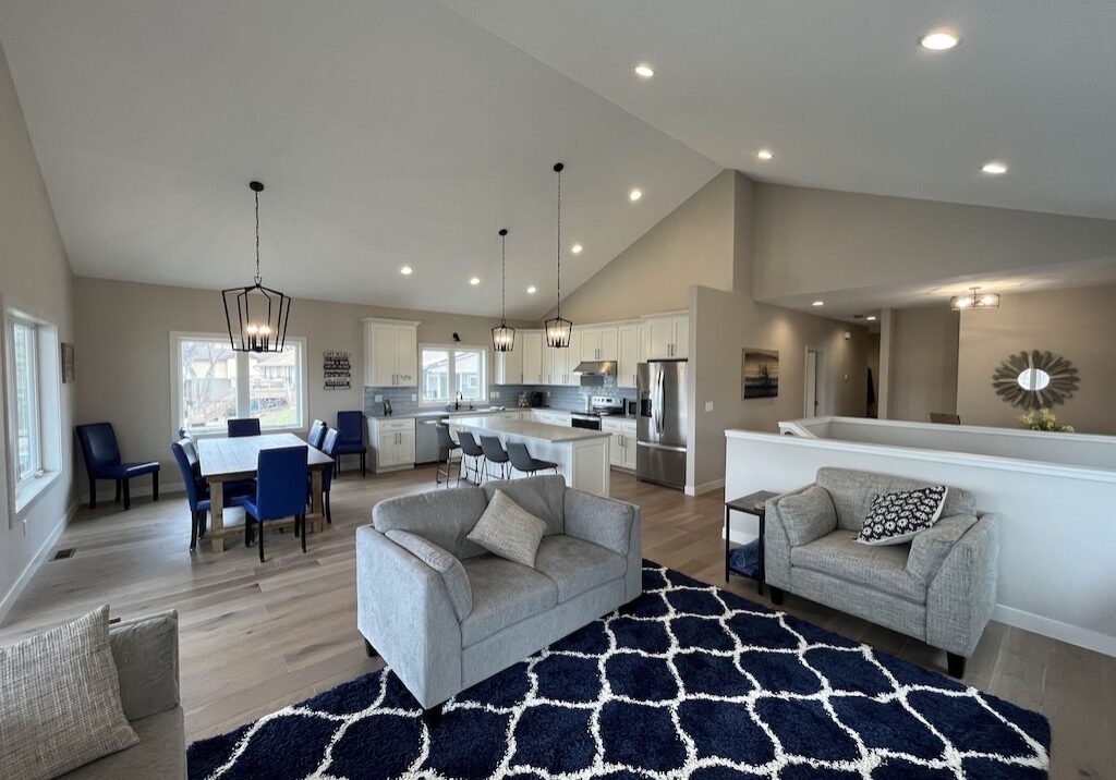 Open-concept living space at Le Homme Dieu Cove with modern kitchen, dining area, and living room. Features gray sofas, a navy geometric rug, blue dining chairs, large windows, pendant lights, and light wood floors.