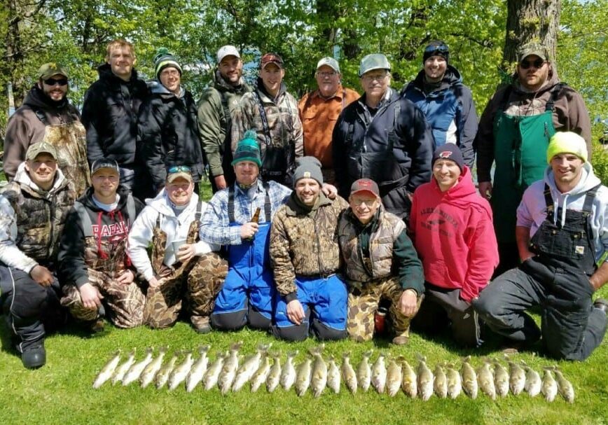 Seventeen people in outdoor clothing pose in two rows on grass, behind a display of freshly caught fish laid out in a row—proof that the 2016 walleye season is off to a great start near Alexandria, MN. Trees and lakeside scenery complete the backdrop.